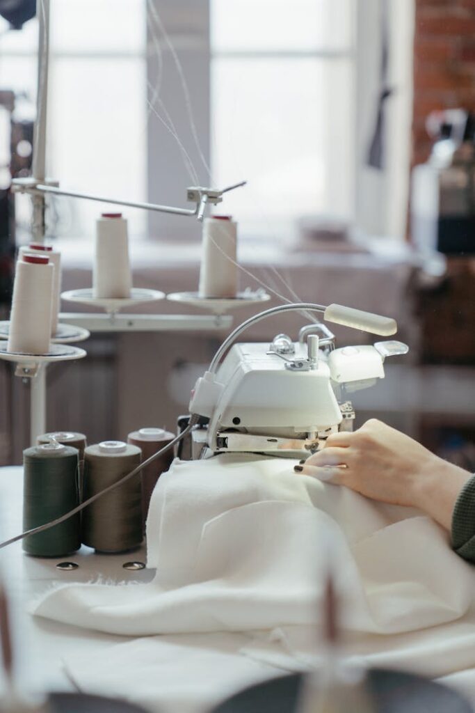 A person sewing fabric on a professional machine in a well-lit workshop with spools of thread.