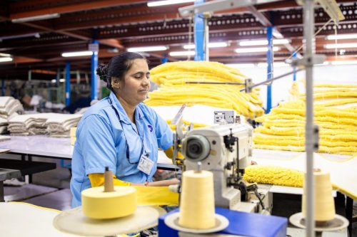 Female textile worker in a factory operating a sewing machine amidst piles of fabric.